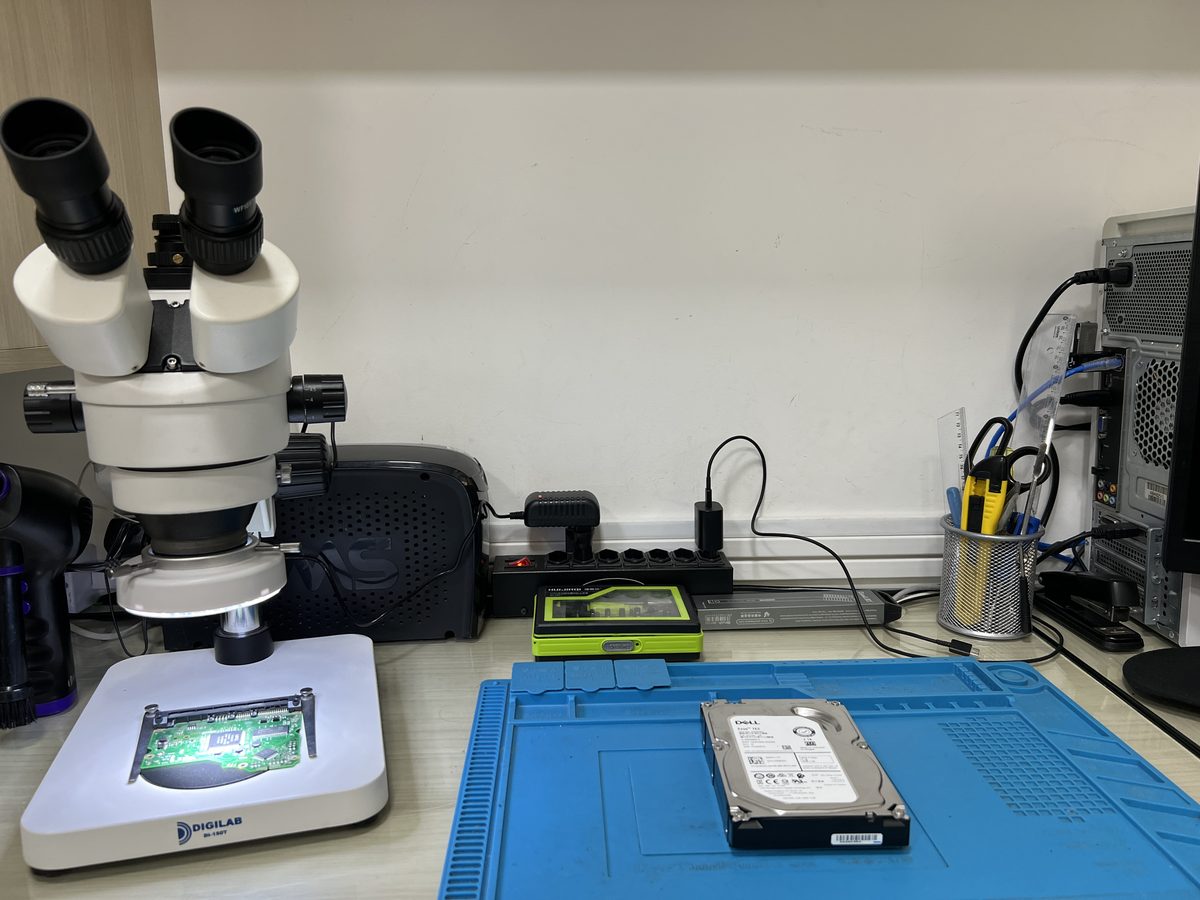 Stereo microscope positioned over a circuit board on an illuminated inspection stage at the Enetsec forensics lab, with a bare hard drive on an ESD mat alongside